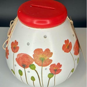 Könnte beinhalten: Weiße Keramik-Sparbüchse mit rotem Deckel und einem Schlitz für Münzen. Die Sparbüchse ist mit roten Mohnblumen und grünen Stielen verziert. Die Blumen haben kleine silberne Glitzerakzente.