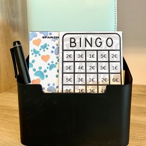 May include: A black organizer with a light blue notebook, a pen, and a bingo card with the text "BINGO" and euro amounts. The bingo card has paw prints and hearts on it.