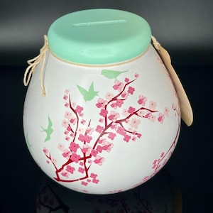 Könnte beinhalten: Weiße Keramik-Sparbüchse mit einem hellgrünen Deckel und einem Kordelverschluss. Die Sparbüchse ist mit einem rosa-weißen Kirschblütenmuster und kleinen grünen Vögeln verziert.