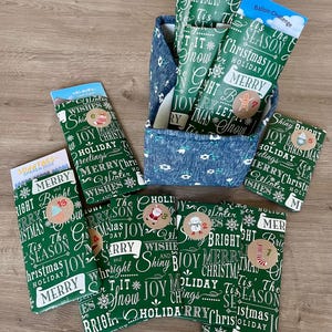 Könnte beinhalten: Eine Auswahl an Weihnachtsgeschenktüten und ein Stoffbehälter. Die Tüten sind grün mit weißem Text und festlichen Illustrationen. Der Behälter ist blau mit einem weißen Blumenmuster. Einige Tüten haben nummerierte Aufkleber.