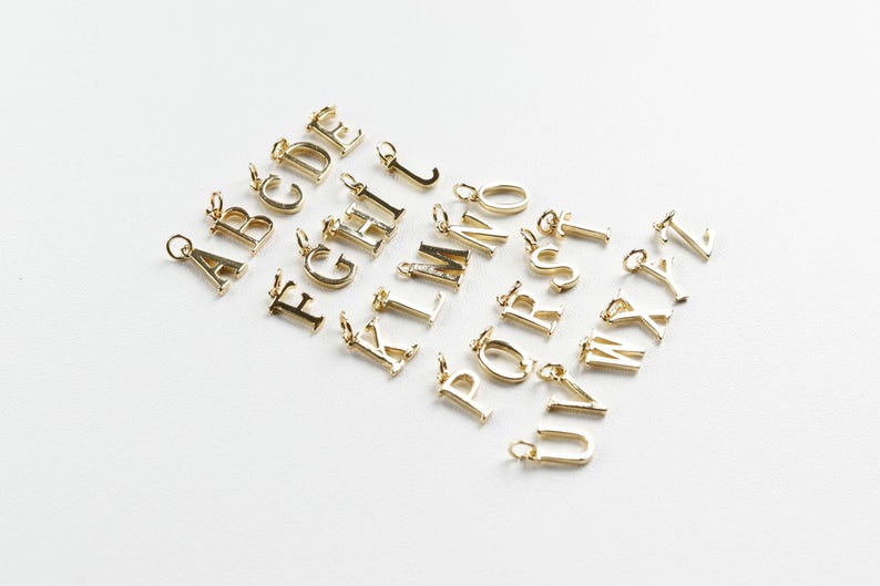 May include: A selection of gold-coloured alphabet charms, each with a small loop for attaching to a chain or bracelet. The letters are in a classic serif font and are scattered on a white surface.