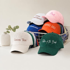 May include: A selection of baseball caps in shades like white, pink, orange, blue, and green. Each cap has embroidered text, including "Bride," "Groomsmen," and "Custom Text." The caps are presented in a white wire basket.