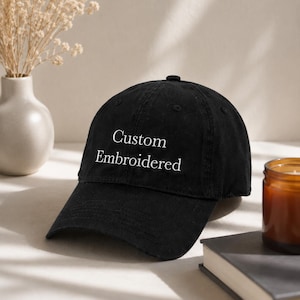 May include: A black baseball cap with the words "Custom Embroidered" in white script. The cap features a curved peak and a rounded crown. The background includes a vase with dried flowers, a candle, and a book.
