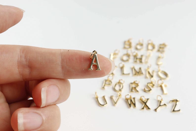 May include: A gold-coloured metal charm in the shape of the letter "A" is held between two fingers. Other gold letter charms are in the background. The charms are likely for jewellery making or personalisation.