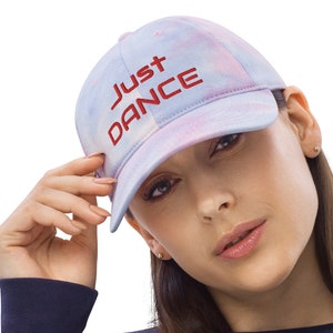 Op de afbeelding: Een blauwe en roze tie-dye baseballpet met de woorden "Just Dance" geborduurd in rood.