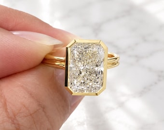 May include: A gold ring featuring a large, rectangular, radiant-cut clear gemstone set in a smooth, polished gold bezel setting. The band consists of two thin, parallel gold wires that meet at the base of the central stone setting.