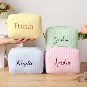 K&ouml;nnte beinhalten: Vier personalisierte Kosmetiktaschen in Gelb, Gr&uuml;n, Blau und Rosa, jeweils mit vertikalem Streifenmuster. Die Namen "Darah", "Sophia", "Kayla" und "Amelia" sind in Schreibschrift auf die Vorderseite gestickt. Jede Tasche hat einen Rei&szlig;verschluss.