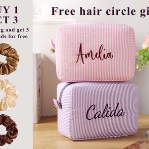 K&ouml;nnte beinhalten: Zwei quadratische Kosmetiktaschen, eine rosa und eine lavendelfarbene, &uuml;bereinander gestapelt. Auf der rosa Tasche ist der Name "Amelia" gestickt, auf der lavendelfarbenen Tasche steht "Calida". Drei Haargummis in Braun- und Cremet&ouml;nen sind ebenfalls zu sehen. Der Text "KAUFE 1 ERHALTE 3" steht oben.