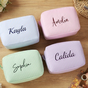 K&ouml;nnte beinhalten: Vier personalisierte Kosmetiktaschen in Pastellfarben: Hellblau, Rosa, Gr&uuml;n und Lavendel. Jede Tasche hat einen anderen Namen in Schreibschrift: Kayla, Amelia, Sophia und Calida. Die Taschen haben eine Seersucker-Textur.