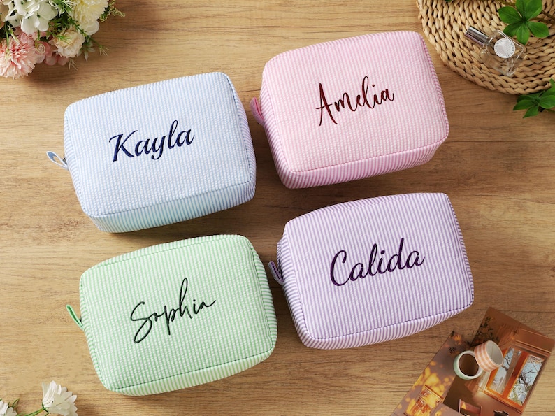 K&ouml;nnte beinhalten: Vier personalisierte Kosmetiktaschen in Pastellfarben: Hellblau, Rosa, Gr&uuml;n und Lavendel. Jede Tasche hat einen anderen Namen in Schreibschrift: Kayla, Amelia, Sophia und Calida. Die Taschen haben eine Seersucker-Textur.