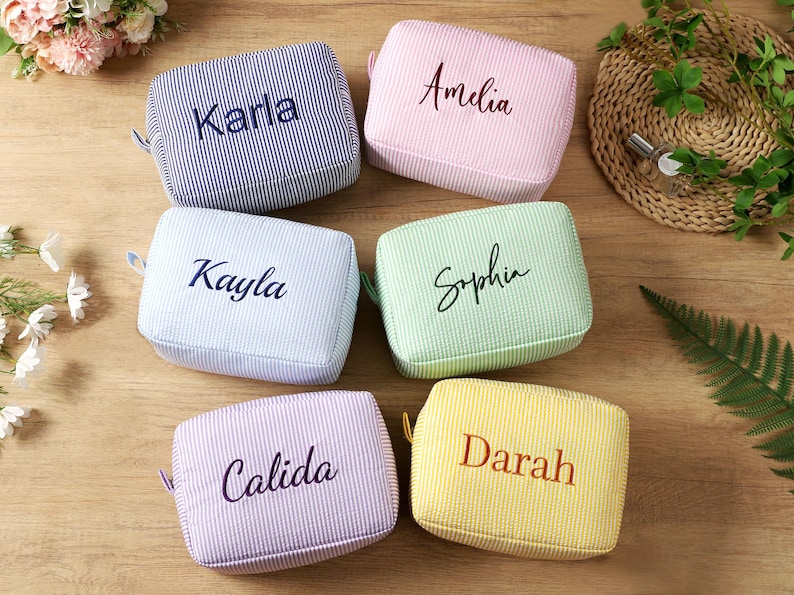 K&ouml;nnte beinhalten: Sechs rechteckige Kosmetiktaschen in Pastellfarben, jede mit einem anderen Namen bestickt. Die Taschen sind gestreift und haben eine kleine Schlaufe. Namen sind Karla, Amelia, Kayla, Sophia, Calida und Darah. Sie liegen auf einer Holzoberfl&auml;che.
