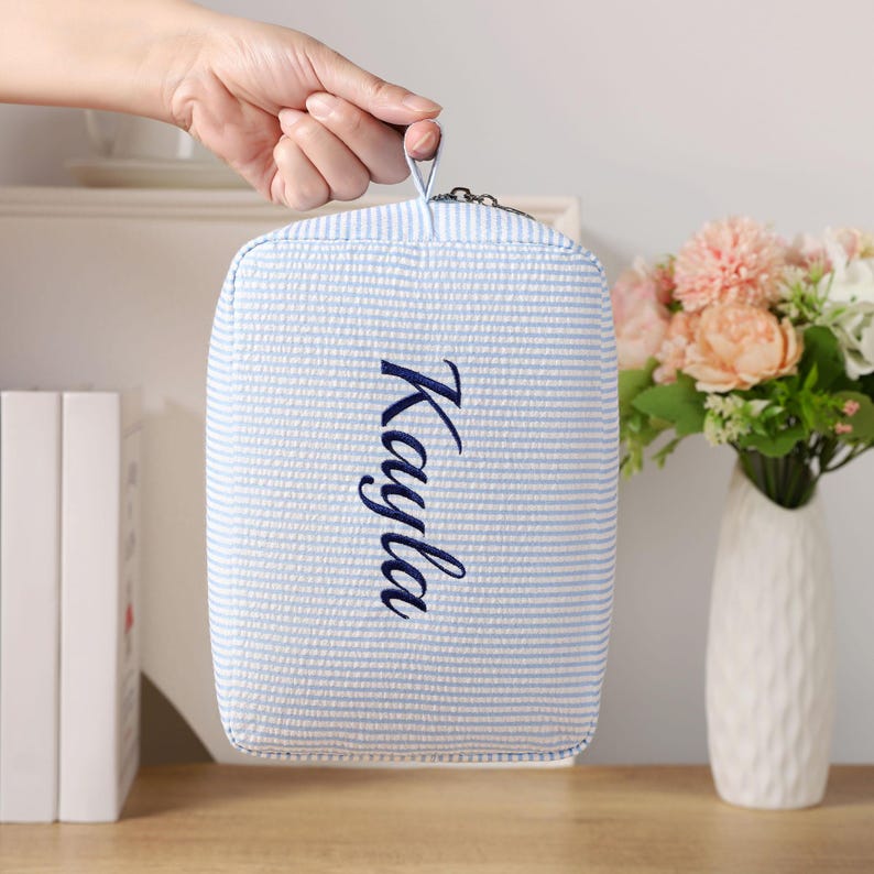 K&ouml;nnte beinhalten: Eine hellblau-wei&szlig; gestreifte Kosmetiktasche mit dem Namen "Kayla" in marineblauer Schrift gestickt. Die Tasche wird von einer Hand gehalten und hat einen kleinen Griff oben. Im Hintergrund steht eine Vase mit Blumen.