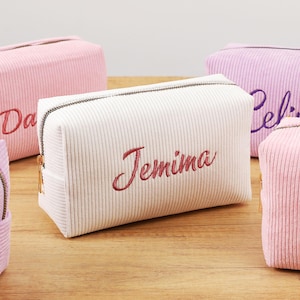 Puede incluir: Varias bolsas de cosméticos de pana en varios colores, incluyendo rosa, blanco y lavanda. Cada bolsa tiene un nombre personalizado bordado en un color contrastante. Las bolsas tienen forma rectangular con cierre de cremallera.