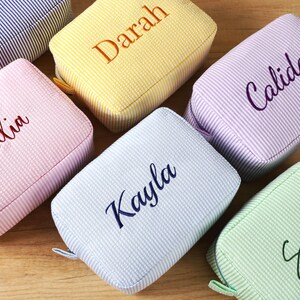 K&ouml;nnte beinhalten: Eine Auswahl an personalisierten Kosmetiktaschen aus Seersucker in Pastellt&ouml;nen wie Rosa, Gelb, Blau und Gr&uuml;n. Jede Tasche ist mit einem Namen in einer passenden Farbe bestickt. Die Taschen sind rechteckig und haben einen Rei&szlig;verschluss.