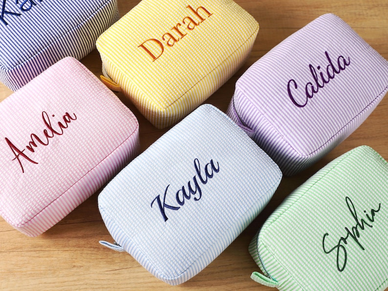 K&ouml;nnte beinhalten: Eine Auswahl an personalisierten Kosmetiktaschen aus Seersucker in Pastellt&ouml;nen wie Rosa, Gelb, Blau und Gr&uuml;n. Jede Tasche ist mit einem Namen in einer passenden Farbe bestickt. Die Taschen sind rechteckig und haben einen Rei&szlig;verschluss.