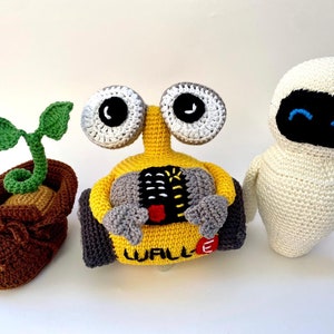 May include: A crocheted set of three toys: a yellow robot with the word "WALL-E" on it, a white robot with blue eyes, and a brown boot with a green plant growing out of it.