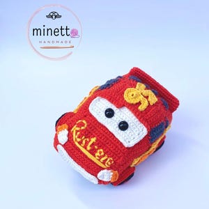 Puede incluir: Un coche rojo de ganchillo hecho a mano con detalles blancos, ojos de botón negros y el texto "Rust-eze". El coche presenta detalles amarillos y azules, y el número 95. El artículo está hecho a mano.
