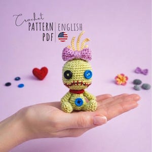 Può includere: Bambola all'uncinetto verde chiaro fatta a mano con un fiocco viola, occhi con bottoni neri e blu e bocca cucita in rosso. La bambola è tenuta in una mano, con il testo "Crochet Pattern English PDF" sullo sfondo.