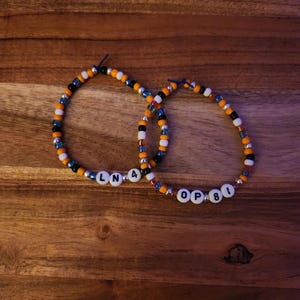 Könnte beinhalten: Zwei Armbänder mit Perlen, auf denen die Buchstaben "LNA4" und "OP81" stehen. Die Armbänder haben eine Mischung aus orangefarbenen, blauen, schwarzen und transparenten Perlen. Die Armbänder werden auf einer Holzoberfläche präsentiert.