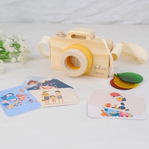 May include: A light-colored wooden toy camera with a yellow lens ring, a white fabric strap, and the text "Amelia's Camera" printed on the side. Included are three translucent colored plastic discs and several rectangular cards with illustrations.