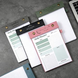 Könnte beinhalten: Eine Sammlung personalisierter Tagesplaner mit Lederbezügen in verschiedenen Farben. Jeder Planer hat Abschnitte für Mahlzeiten, To-Do-Listen und Notizen. Die Einbände sind mit Namen und Titeln geprägt und mit messingfarbenen Druckknöpfen befestigt.