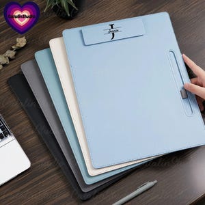 Könnte beinhalten: Eine Auswahl an Leder-Klemmbrettern in Schwarz, Grau, Hellblau und Creme. Das oberste Klemmbrett ist hellblau und trägt den Namen "Jennie" und ein stilisiertes "J". Ein Stift ist im Vordergrund zu sehen. Die Klemmbretter sind gestapelt.