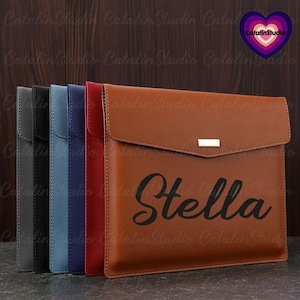 May include: A brown leather laptop sleeve with a flap closure and a gold metal accent. The sleeve has the name "Stella" engraved in black lettering.