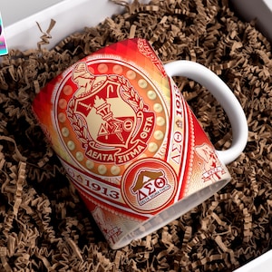 May include: A white ceramic mug with a red and gold design featuring Greek letters and the text "Delta Sigma Theta 1913." The mug is inside a white box filled with brown crinkle paper.