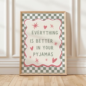 May include: A framed print with the text "EVERYTHING IS BETTER IN YOUR PYJAMAS". The text is in a white, wavy-edged shape, with pink accents and decorative stars and hearts, set against a green and white checkered background.