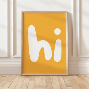 Peut inclure: Une affiche encadrée rectangulaire présentant un fond jaune moutarde avec le mot hi écrit en grandes lettres minuscules blanches, grasses et arrondies, centrées au milieu du cadre. Le cadre est fabriqué dans un matériau en bois clair.