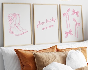 May include: Three framed prints with pink illustrations. The left print shows a pair of pink cowboy boots. The middle print has the text "How lucky are we". The right print shows five pink bows.