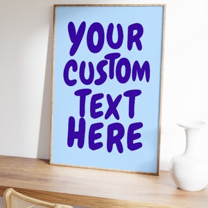 May include: A framed poster with a light blue background and the text "YOUR CUSTOM TEXT HERE" in bold, purple letters. The poster is sitting on a wooden desk with a white vase.