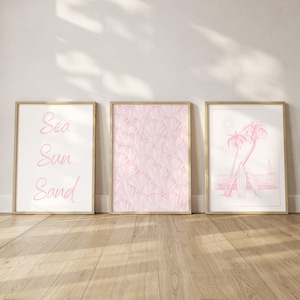 May include: Three framed art prints with a beach theme. The left print has the words "Sea Sun Sand" in pink script. The middle print has a pink and white abstract pattern. The right print features a pink illustration of palm trees, surfboards, and the ocean.