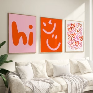 Può includere: Tre stampe incorniciate con uno sfondo rosa e testo arancione che dice "hi", uno sfondo rosso con faccine sorridenti rosa e uno sfondo bianco con cuori rosa.