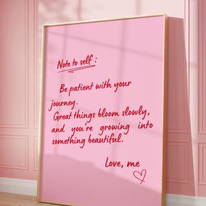 May include: A pink framed print with the text "Note to self: Be patient with your journey. Great things bloom slowly, and you're growing into something beautiful. Love, me" in red script. The print is leaning against a pink wall.