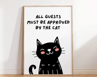 May include: A framed art print with the text "ALL GUESTS MUST BE APPROVED BY THE CAT" above a black cartoon cat with pink cheeks. The print is in a light wood frame and is displayed on a white wall.
