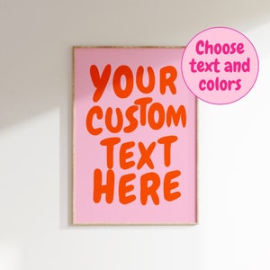 May include: A pink framed poster with the text "YOUR CUSTOM TEXT HERE" in bold, orange letters. The text is centered on the poster. The poster is on a white wall.