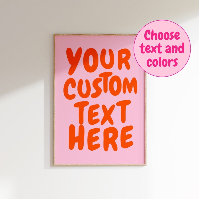 May include: A pink framed poster with the text "YOUR CUSTOM TEXT HERE" in bold, orange letters. The text is centered on the poster. The poster is on a white wall.