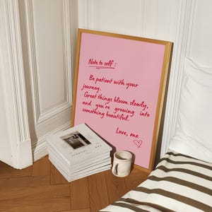 May include: A pink framed print with the text "Note to self: Be patient with your journey. Great things bloom slowly, and you're growing into something beautiful. Love, me." A stack of books and a small mug are in front of the print.