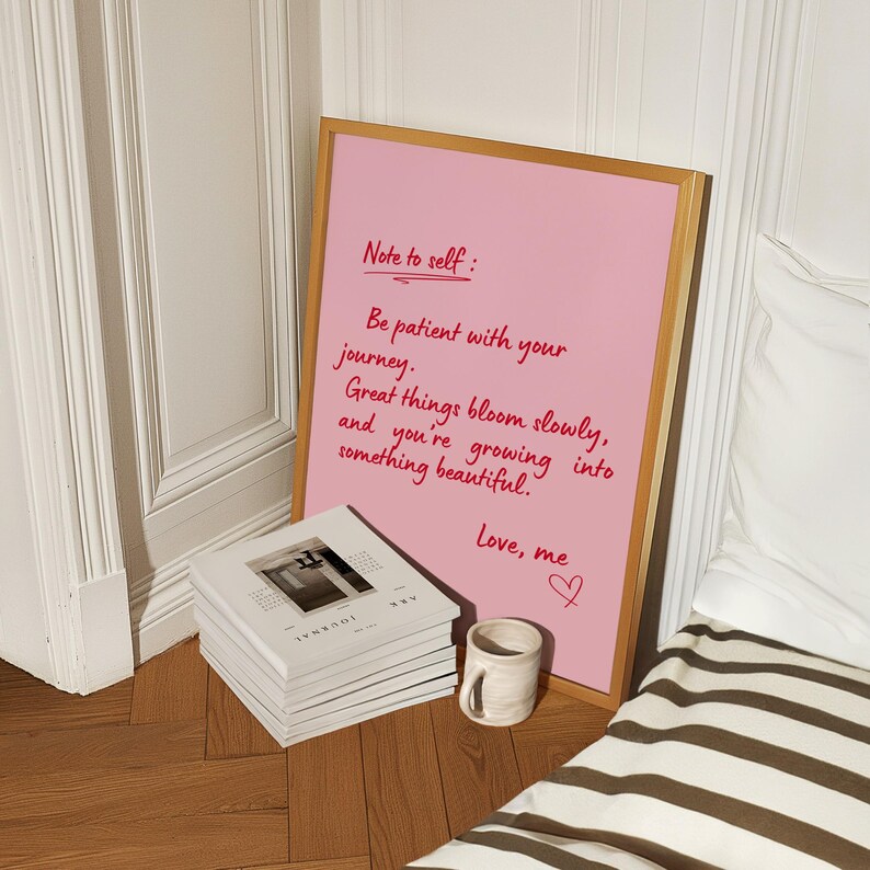 May include: A pink framed print with the text "Note to self: Be patient with your journey. Great things bloom slowly, and you're growing into something beautiful. Love, me." A stack of books and a small mug are in front of the print.