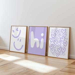 Peut inclure: Trois cadres muraux rectangulaires. L'affiche de gauche présente deux visages souriants violet clair sur fond blanc. L'affiche centrale affiche le texte blanc "hi" sur fond violet clair. L'affiche de droite montre un motif de cœurs violet clair sur fond blanc.