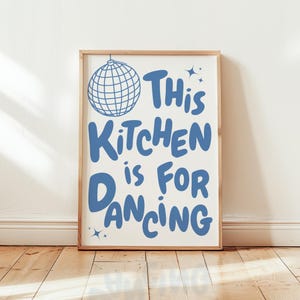 以下が含まれることがあります： 「This Kitchen is For Dancing」という青い文字と、ディスコボールのイラストが描かれた額入りのプリント。白い背景に、明るい茶色の木製フレームで額装されています。プリントは、木製の床に白い壁に立てかけられています。