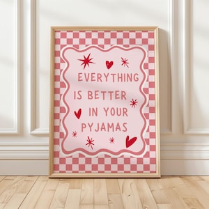 以下が含まれることがあります： 「EVERYTHING IS BETTER IN YOUR PYJAMAS」のテキストが入った額入りのプリント。ピンクと赤の配色で、ハートとスターバーストがチェック柄の背景に配置されています。フレームは明るい木の色です。寝室に最適です。