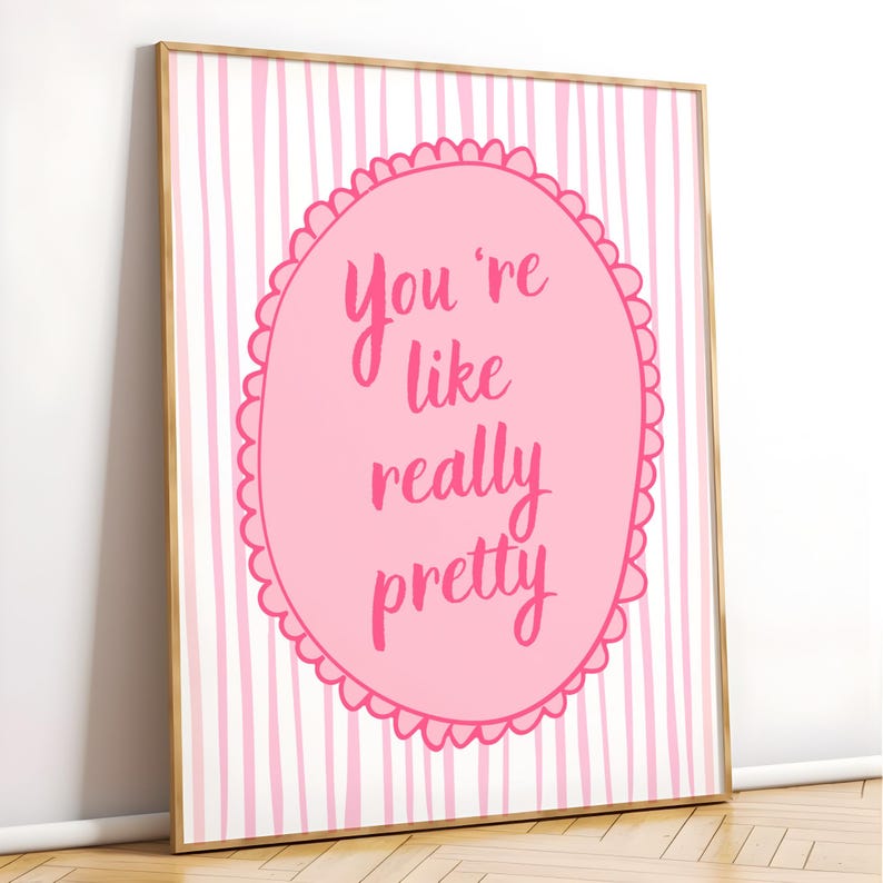 May include: A framed art print with a pink and white striped background. A pink oval in the center features the handwritten text "You're like really pretty" in a darker pink. The frame is a light gold color.