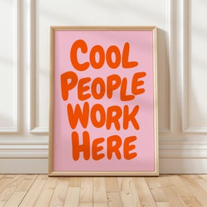 Op de afbeelding: Een ingelijste print met de tekst "COOL PEOPLE WORK HERE" in oranje letters op een roze achtergrond. De lijst is licht van hout, en de print wordt gepresenteerd tegen een witte muur en een houten vloer.