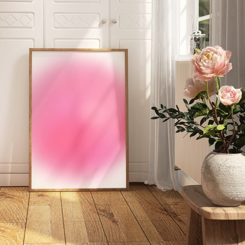 Puede incluir: Una pintura abstracta enmarcada con un degradado rosa y blanco. La pintura est&aacute; colgada en una pared blanca en una habitaci&oacute;n con suelo de madera y un jarr&oacute;n de flores rosas.