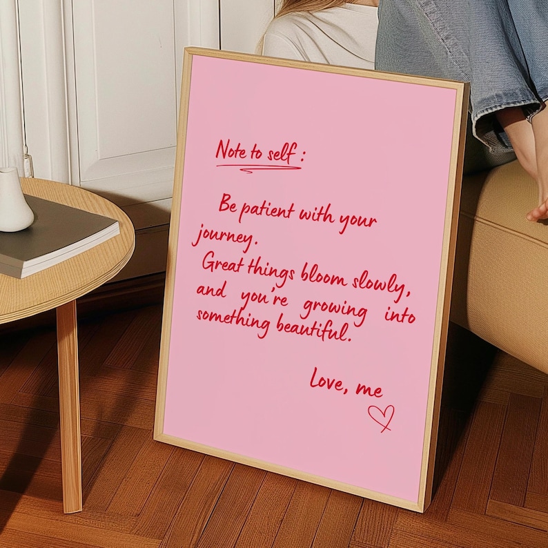 May include: A pink framed sign with the text "Note to self: Be patient with your journey. Great things bloom slowly, and you're growing into something beautiful. Love, me" written in red script.
