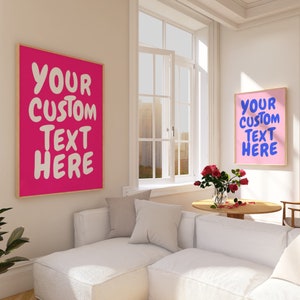 May include: Two framed prints with pink and light pink backgrounds and white text that reads "YOUR CUSTOM TEXT HERE" in a playful, handwritten font. The prints are hanging on a white wall in a living room setting with a white couch and a small table with a vase of pink flowers.