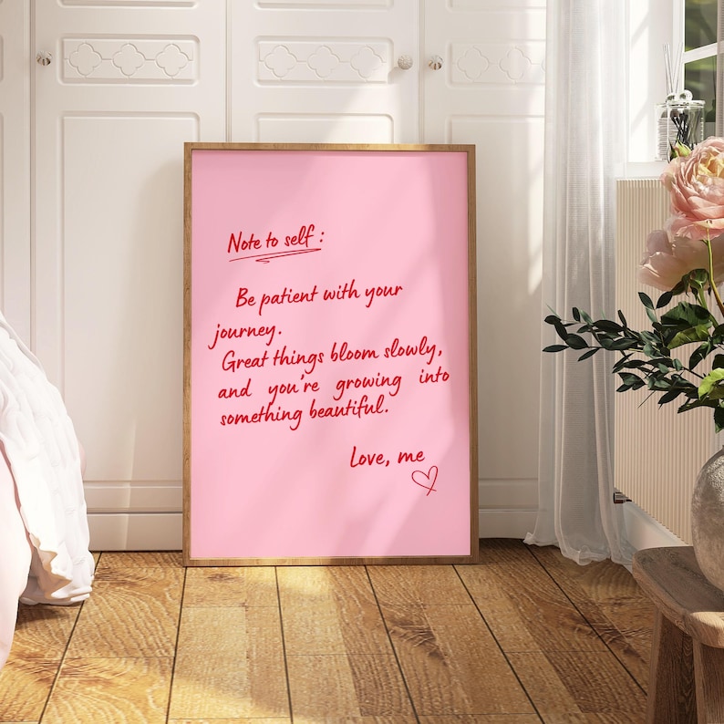 May include: A framed pink print with the text "Note to self: Be patient with your journey. Great things bloom slowly, and you're growing into something beautiful. Love, me." The frame is a light brown color.