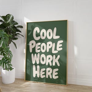 Puede incluir: Un póster enmarcado con el texto "COOL PEOPLE WORK HERE" en letras blancas, en negrita y redondeadas sobre un fondo verde oscuro. El marco es dorado. Una planta en maceta está a la izquierda del póster.